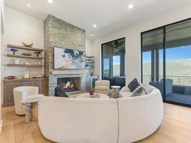 Stone-clad fireplace with a stone mantel, wood-finish flooring, built-in shelving, and large sliding glass doors leading to an outdoor deck