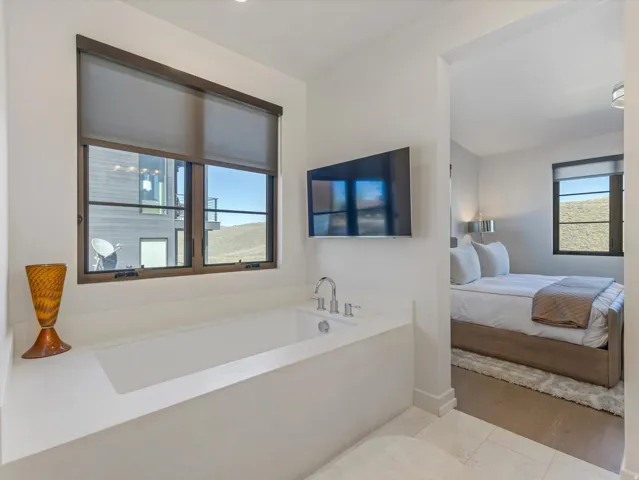 Built-in soaking tub with deck-mounted faucet, large window with roller shade, and flat-panel television