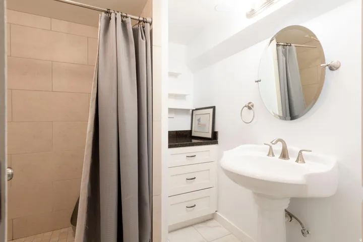 Shower with tiled walls, pedestal sink, oval mirror, white cabinetry, and a dark stone countertop