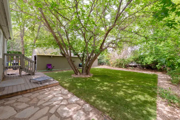 Expansive backyard featuring a manicured lawn, flagstone patio, wood deck with railings, and mature trees providing natural shade