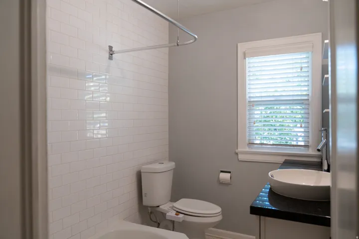 White subway tile shower surround, window with white blinds, dark stone countertop, white vessel sink, and chrome fixtures