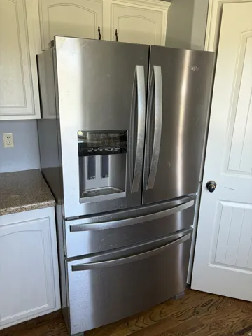 Stainless steel French door refrigerator with bottom freezer, integrated ice and water dispenser, framed by white cabinetry, and a light-colored countertop