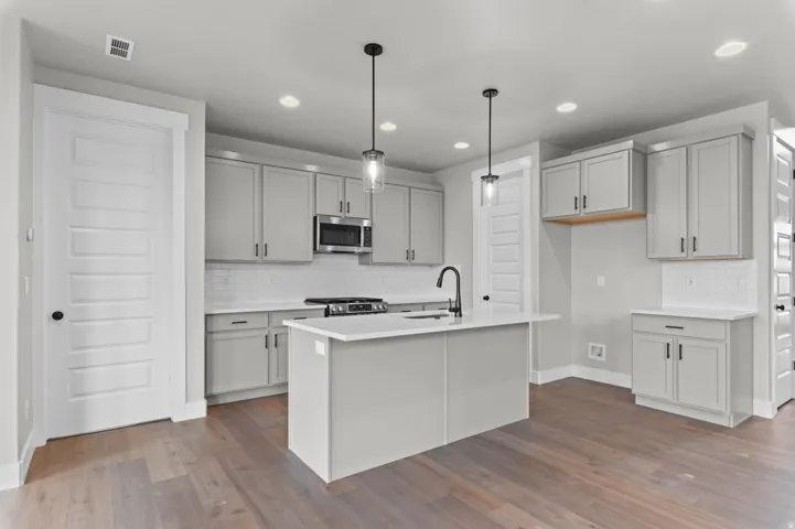 Kitchen featuring wood-finish flooring, an island with a built-in sink, stainless steel microwave, gray cabinetry, and pendant lighting