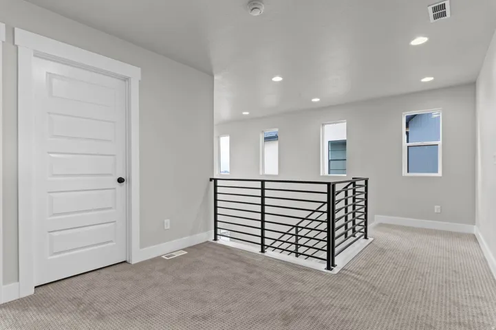 Light-toned carpeting, modern black horizontal bar railing, contemporary white paneled door, multiple recessed ceiling lights, and multiple windows