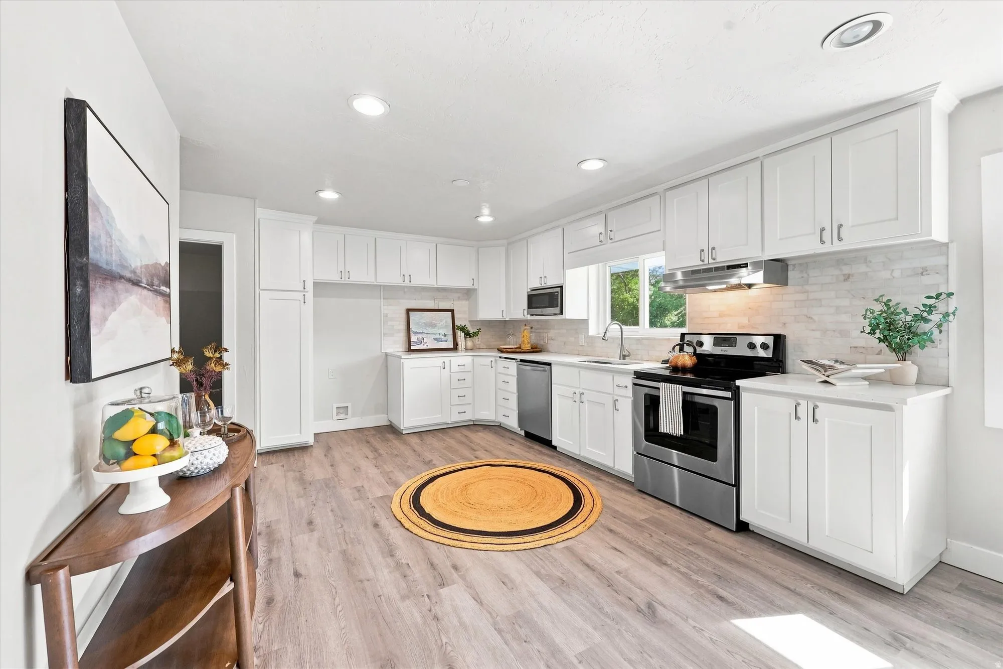 White shaker cabinetry, stainless steel appliances, wood-finish flooring, recessed lighting, and a window over the sink