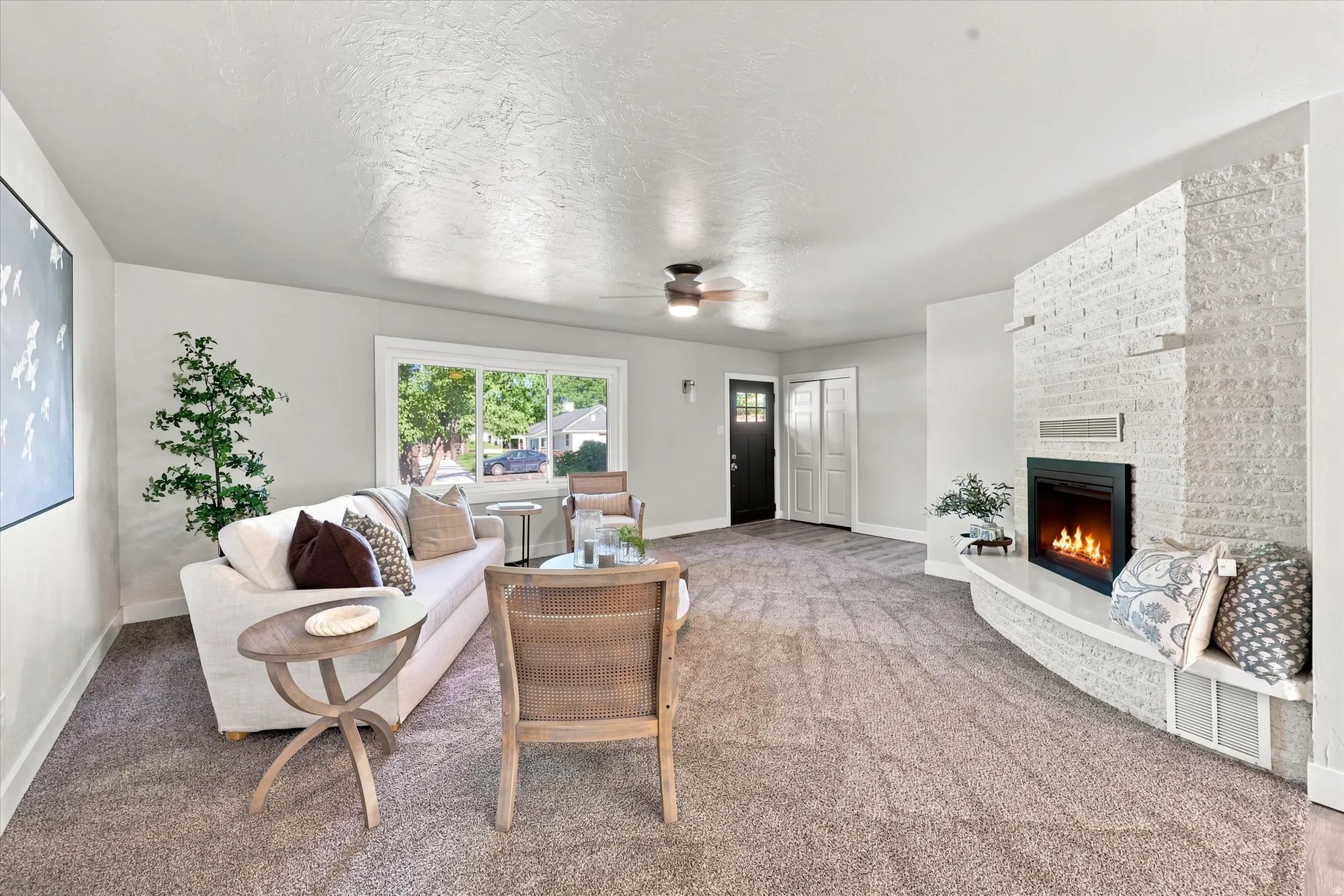 Spacious living area featuring carpet flooring, a white-painted brick fireplace with a hearth, a ceiling fan, and large windows