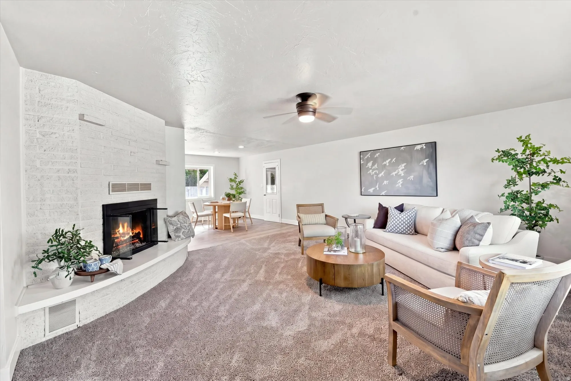 White-painted brick fireplace with a hearth, gray carpeting, a ceiling fan, and a dining area with a window