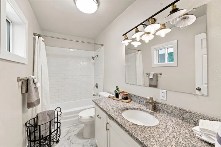 Bathroom featuring a white subway tile shower surround, a built-in bathtub, a window, a single vanity with a stone countertop, and a mirror with a multi-bulb light fixture