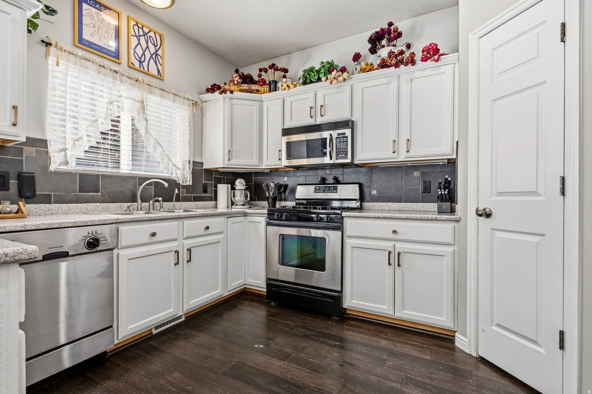 White cabinetry with dark hardware, stainless steel appliances, dark tile backsplash, light-colored countertops, and wood-finish flooring