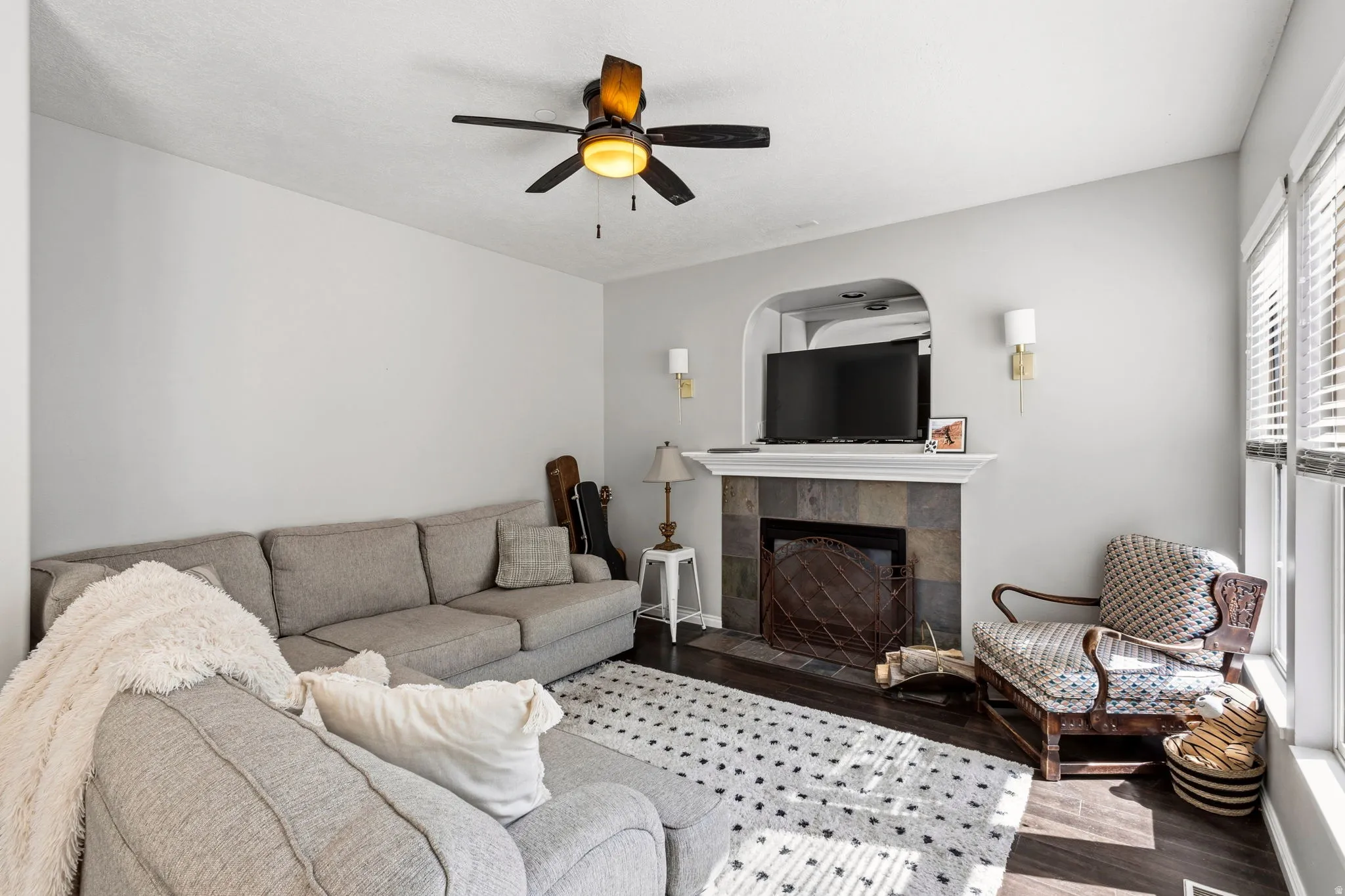 Recessed fireplace with stone surround, dark wood-finish flooring, ceiling fan with integrated lighting, and multiple windows with blinds