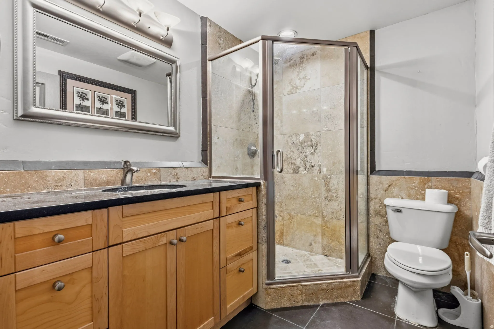 Vanity with a basin sink and a dark countertop, wood-finish cabinetry, corner glass shower enclosure with stone-finish tile surround, and stone-finish tile wainscoting