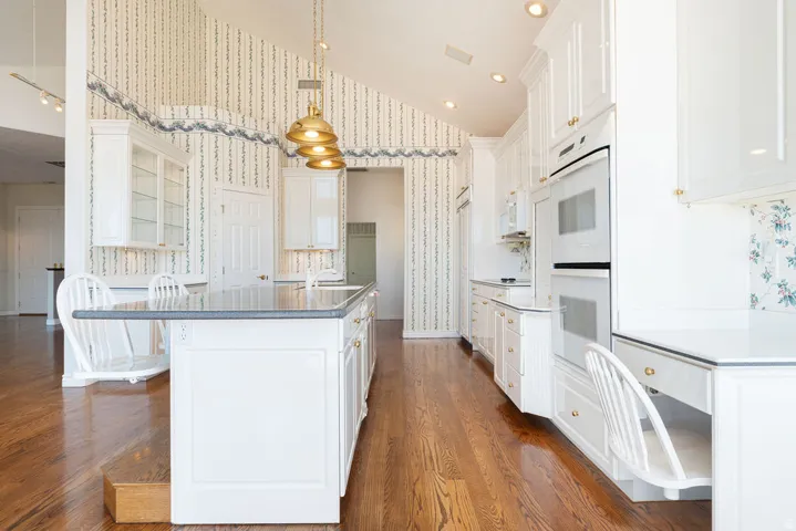 Vaulted ceiling, wood-finish flooring, white cabinetry, integrated double ovens, and a kitchen island with a dark countertop
