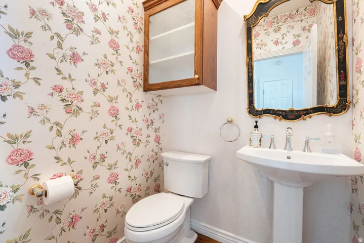 Pedestal sink with chrome fixtures, an ornate framed mirror, a wall-mounted wood-finish cabinet, and floral wallpaper