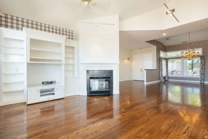 Hardwood-finish flooring throughout, built-in media center with shelving, white mantel fireplace with dark stone surround, vaulted ceilings, and a bay window with a chandelier