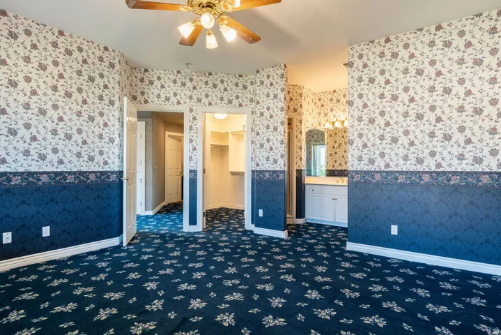 Ceiling fan with light fixture, patterned carpeting, dual-tone wallpaper, built-in shelving, and a vanity with a mirror