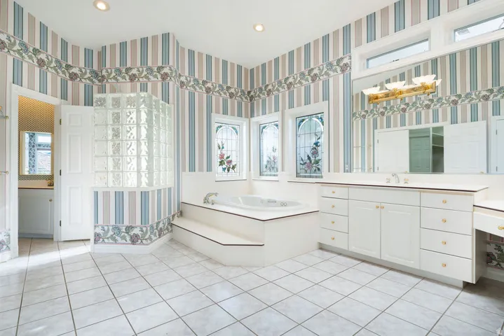 White tile flooring, built-in soaking tub, dual white vanities, and stained-glass window accents