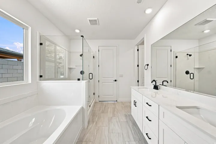 Recessed lighting illuminates a spacious bathroom featuring a white soaking tub, glass-enclosed shower, and a dual-sink vanity with white cabinetry and dark hardware