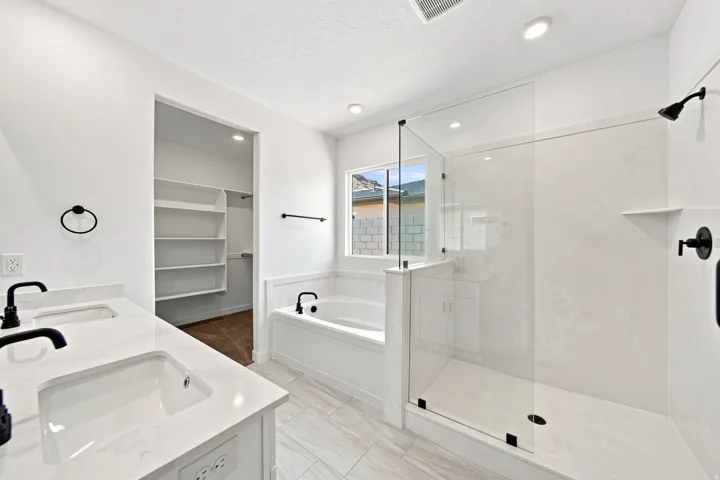 Dual vanity with undermount sinks and black fixtures, a built-in soaking tub, a glass-enclosed shower, a built-in closet with shelving, and light-toned tile flooring