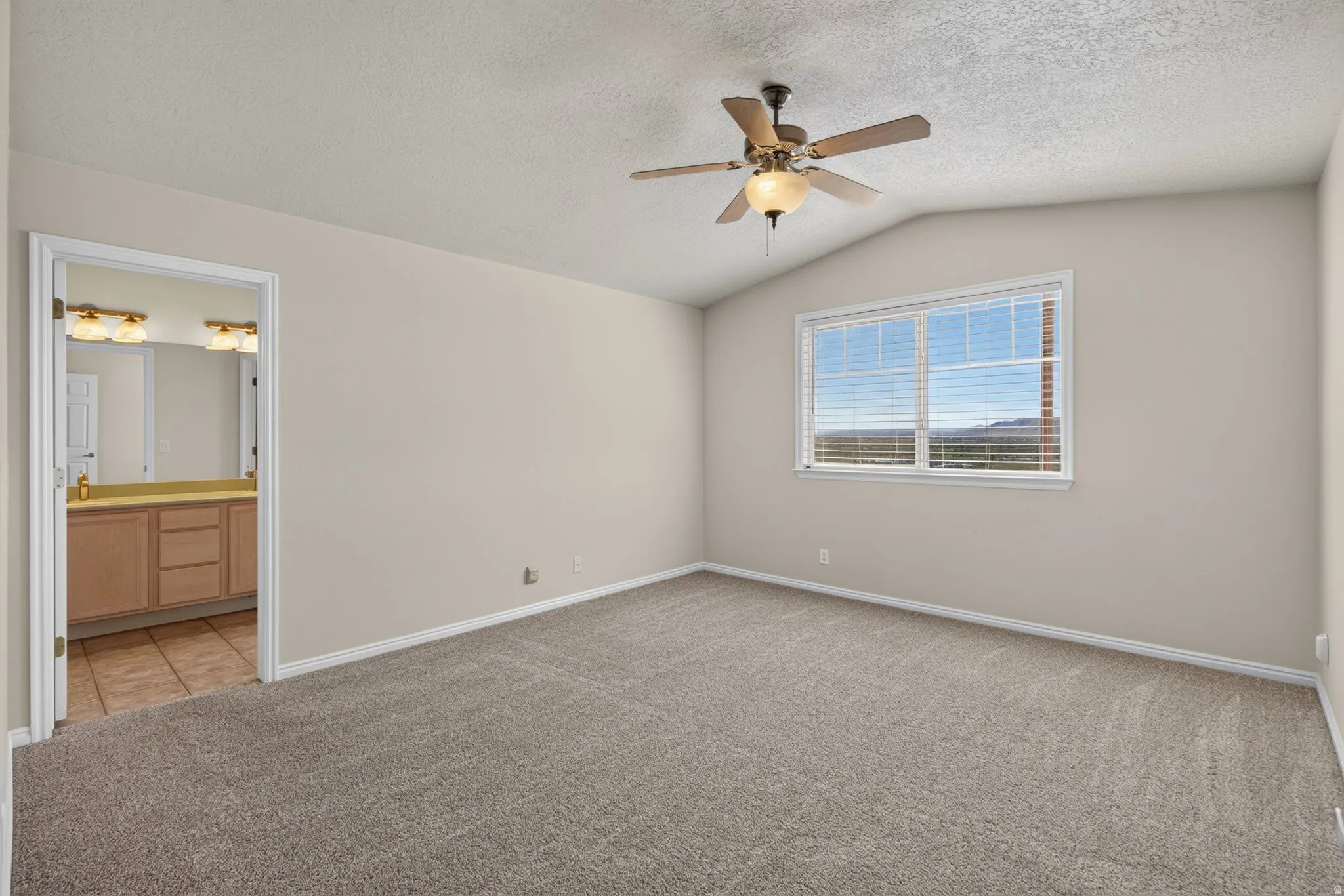 Vaulted ceiling, ceiling fan with integrated lighting, large window with blinds, neutral carpet, and a bathroom vanity with dual sinks