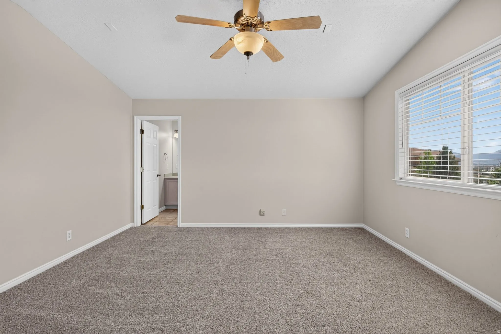 Spacious room featuring neutral carpeting and wall tones, a ceiling fan with integrated lighting, and a large window with blinds