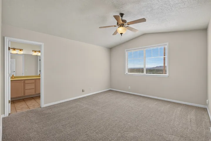 Vaulted ceiling, ceiling fan with integrated lighting, large window with blinds, neutral carpet, and a bathroom vanity with dual sinks