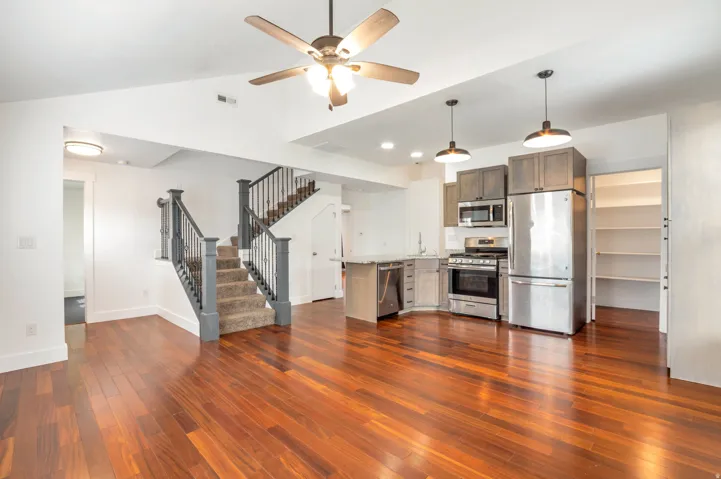 Open-concept living area featuring rich wood-finish flooring, a contemporary kitchen with stainless steel appliances, and a staircase with dark wood newel posts and railing