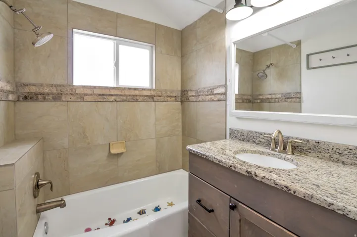 Bathroom featuring a tub/shower combination with a built-in bench, tiled walls with a decorative accent band, a window, a single vanity with a granite-style countertop, and an integrated sink with a brushed nickel faucet