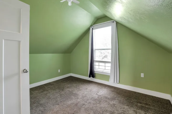Sloped ceiling room featuring a window with blinds, carpet flooring, a white interior door, and a ceiling fan