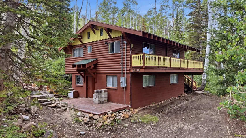 Log cabin exterior with prominent wood siding, upper-level balcony with yellow railing, covered entryway, and a ground-level deck