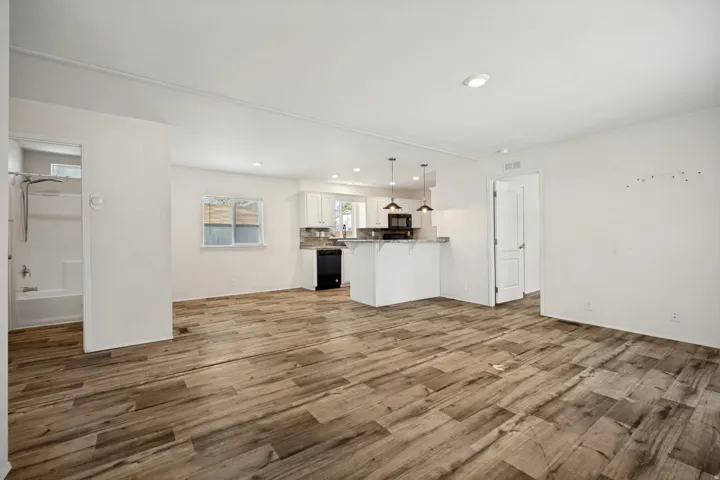 Spacious open-concept living area with wood-finish flooring, white walls, recessed lighting, and a kitchen featuring white cabinetry, black appliances, and pendant lighting