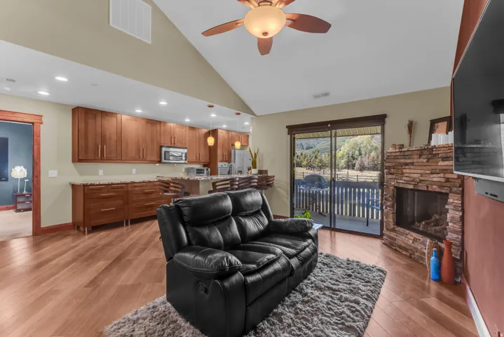 Vaulted ceiling with a ceiling fan, wood-finish flooring, stone-clad fireplace, sliding glass door to exterior, and wood cabinetry with stainless steel appliances