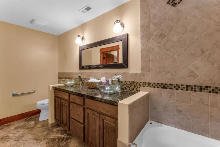 Dual glass vessel sinks set on a dark countertop, wood-finish cabinetry, tile flooring, and a tiled shower surround with a decorative mosaic band