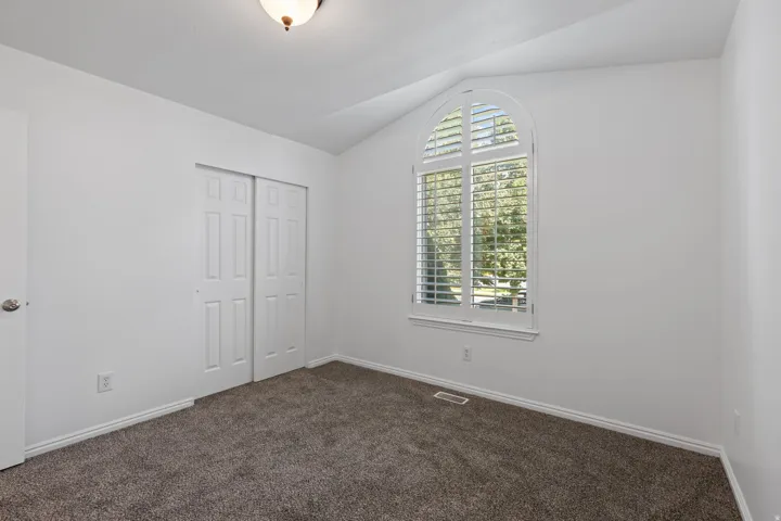 Arched window with plantation shutters, vaulted ceiling, neutral wall color, carpeted flooring, and sliding panel closet doors