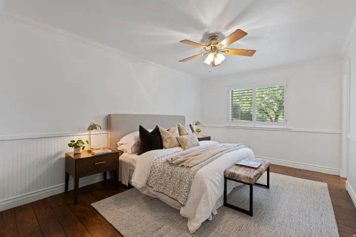 Hardwood flooring, crown molding, white wainscoting, wood-blade ceiling fan, and window with plantation shutters