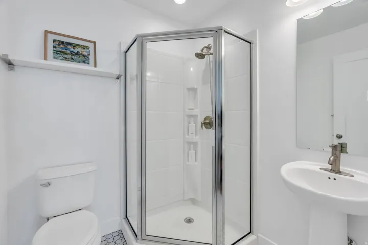 Pedestal sink with brushed nickel faucet, walk-in shower with clear glass enclosure and built-in shelving, white hexagonal floor tiling, and overhead lighting