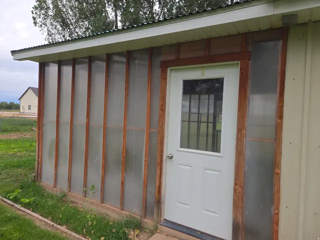 Enclosed structure featuring a white entry door with a window, corrugated metal roof, and translucent wall panels with wood framing