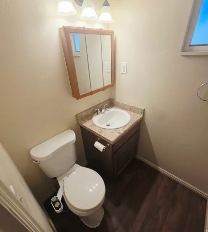 Wood-finish flooring, single vanity with integrated sink, wood-finish cabinet, wall-mounted light fixture, and window
