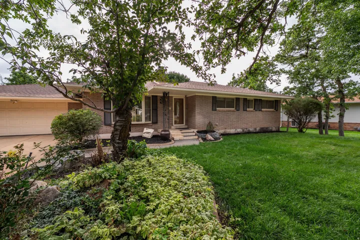 Brick exterior with contrasting dark window shutters, an attached garage, a paved driveway, and established front landscaping
