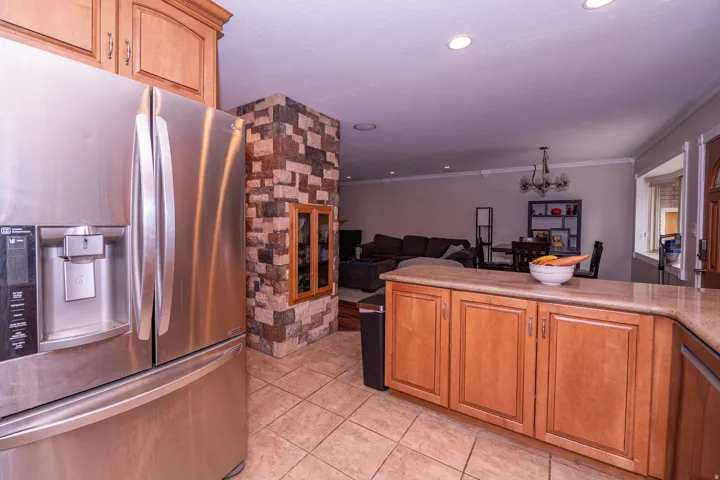 Stainless steel refrigerator, light wood-finish cabinetry, light-toned countertop, tiled flooring, and a stone-clad architectural feature