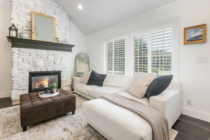 Vaulted ceiling, stone-clad fireplace with mantel, dark wood-finish flooring, and white plantation shutters on large windows