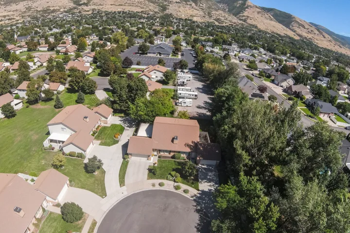 Aerial view featuring residential properties with tan roofs, ample green landscaping, and mature trees, set against a mountainous backdrop
