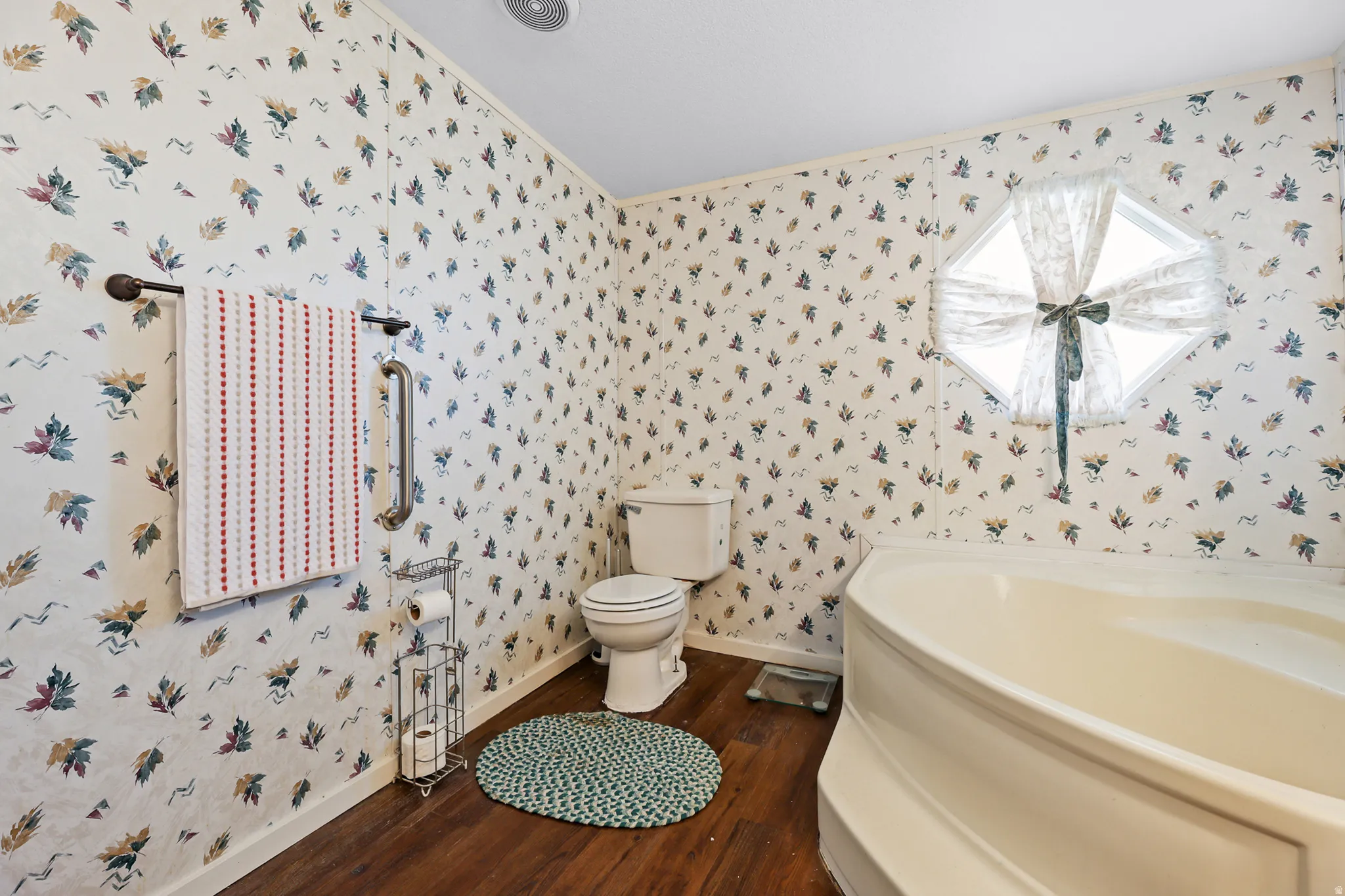 Soaking tub with a unique octagonal window, wood-finish flooring, and decorative wallpaper