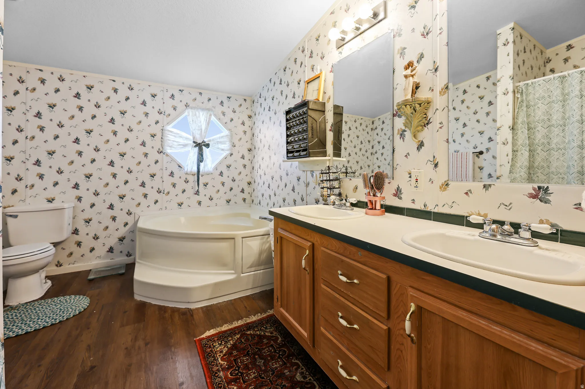 Dual vanity with integrated sinks, wood-finish cabinetry, built-in corner bathtub, and wood-finish flooring