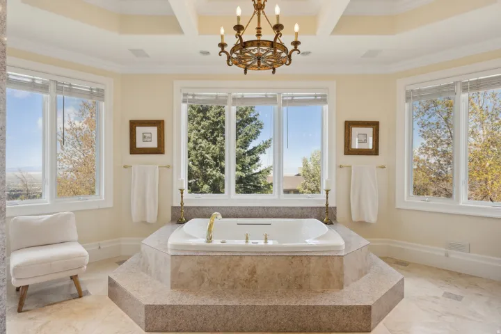 Recessed soaking tub with a stone-clad surround, tray ceiling with chandelier, and multiple windows