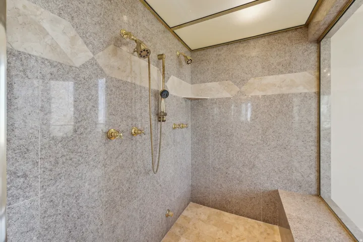 Walk-in shower with stone tile walls, built-in bench, and multiple brass shower fixtures