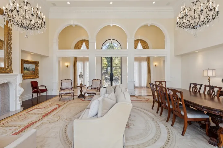 Double-height ceiling with decorative molding, grand chandeliers, arched doorways, dark wood-finish flooring, and a carved stone fireplace