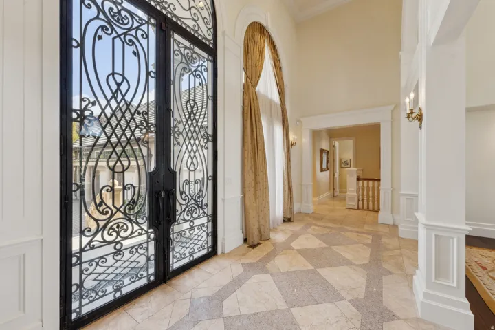Wrought iron and glass double entry doors, checkerboard pattern flooring, tall archway with drapery, and decorative wall sconces