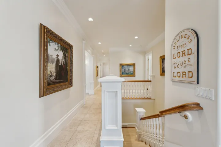 Elegant second-floor hallway featuring recessed ceiling lighting, decorative framed artwork, white painted wainscoting, wood-finish handrail, and neutral wood-finish flooring throughout