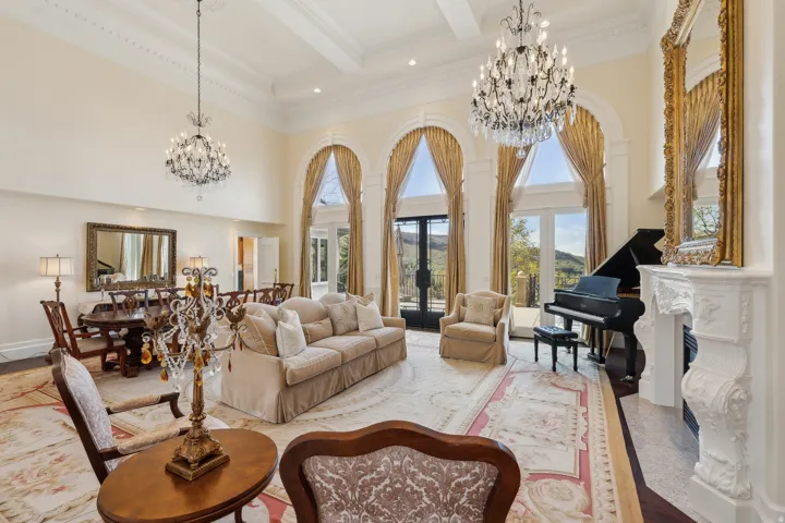 Grand living area featuring vaulted ceilings, multiple crystal chandeliers, arched windows with exterior scenery, and a decorative fireplace with a carved mantel
