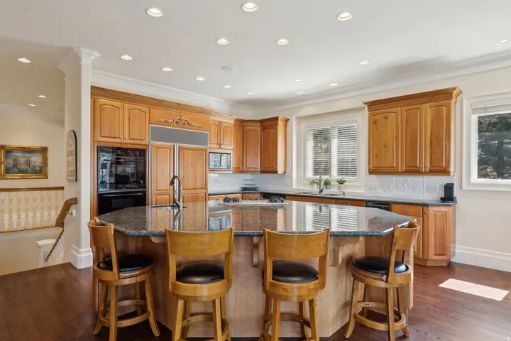 Spacious kitchen featuring wood-finish cabinetry, a large center island with counter seating, built-in appliances, dark stone countertops, and recessed lighting